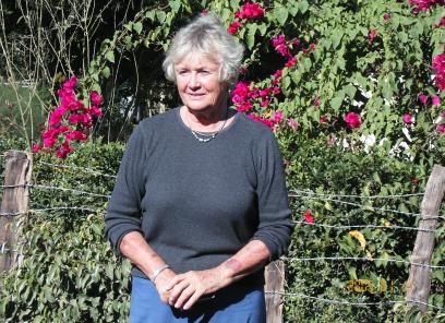 An elderly white woman with short white hair stands in front of lush greenery and pink flowers in Haiti. She wears a black sweater and clasps her hands at her waist.