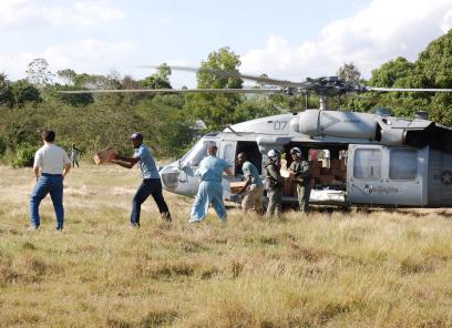 A helicopter sits in a wide field with trees in the background. Doctors and staff memebrs of a hospital unload supplies from the helicopter in an assembly line. 