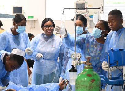 A half-dozen Haitian clinicians in blue medical gowns surround the bed of an ER patient who is receiving IV fluids.
