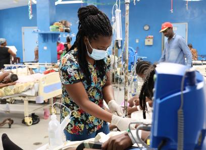 An woman Haitian emergency room clinician tends to the patient. The patient is mostly hidden behind a blue patient monitor. The clinician has long braids and wears a patterend scrub top, a blue mask and white exam gloves.