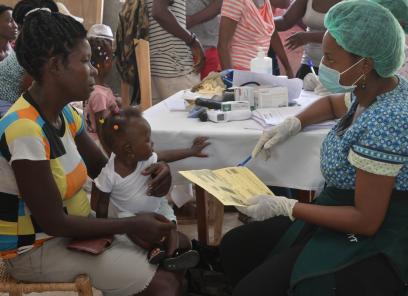 A Haitian woman holding a baby on her lap speaks with a nurse in face mask and gloves. They sit at a table, apart from a crowd.