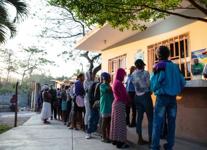 In early-morning light, a line of Haitian men and women stretches from a pale yellow building out through open hospital gates.