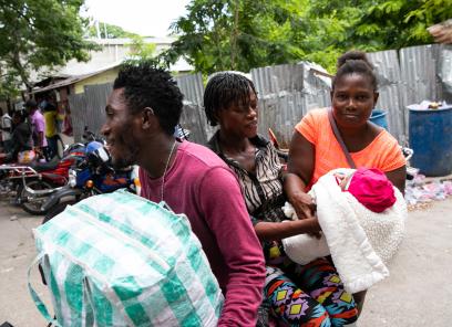 On a street corner in Haiti, two women hold an infant wrapped in a blanket. A man carrying a large tote bag passes next to them.