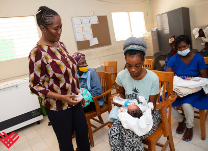 A Haitian clinician wearing a maroon, white, and black patterned shirt talks with a young mother who is sitting in a chair cradling her baby. 