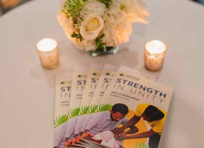 A round tabletop holding a bouquet of white flowers, 2 candles, and a stack of programs with the words “Strength in Unity” and a cutout photo of an infant being vaccinated.