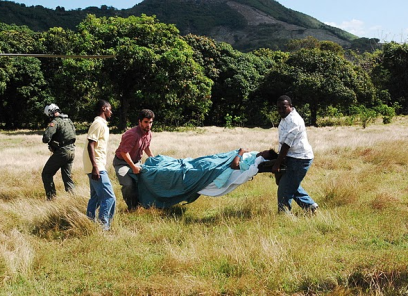 Three men carry a stretcher with a patient on it across a grassy field.
