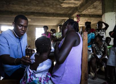 A male Haitian doctor examines two patients at a crowded community health clinic.