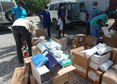 A group of people unpack cardboard boxes full of medical supplies. Some of the boxes are open and you can see blue medical masks and packages wrapped in a white wrapper. A man in black jeans and a yellow polo shirt bends down to unpack the boxes. Nurses in blue t-shirts in the background observe. There is a white ambulance with green lettering in the background. The ground is white and grey gravel.