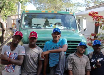 A group of five Haitian men sit in front of a large, light blue truck. They are all smiling and wearing blue or red baseball caps.