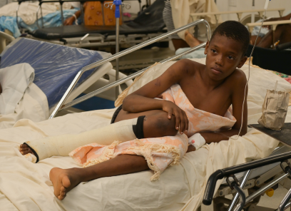 A young boy lies in a hospital bed with a leg injury. He is covered by an orange and white blanket. His right leg is wrapped in a bandage below the knee.