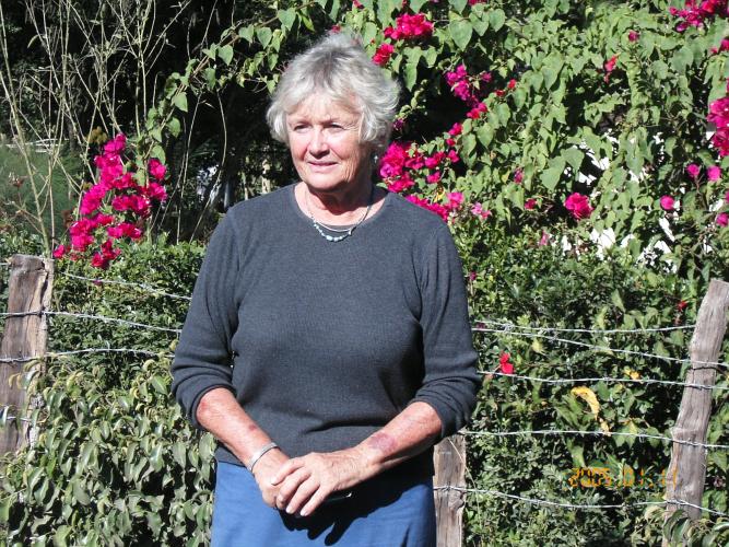 An elderly white woman with short white hair stands in front of lush greenery and pink flowers in Haiti. She wears a black sweater and clasps her hands at her waist.