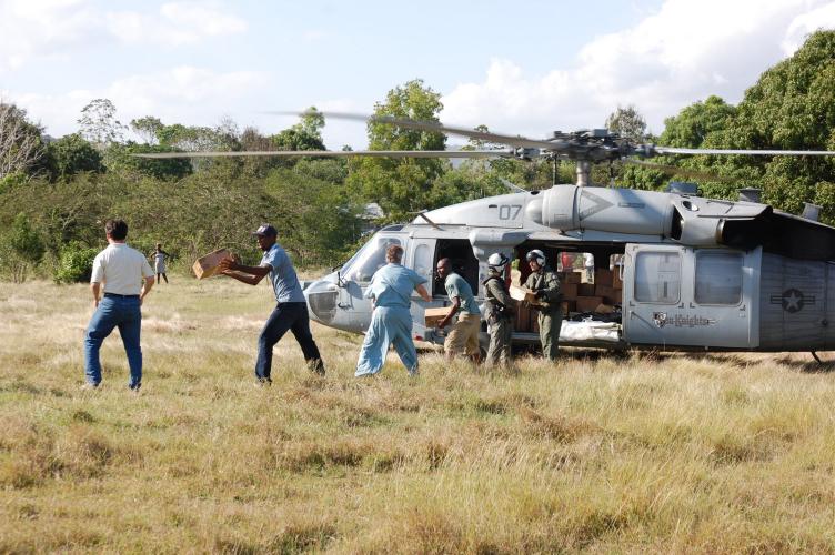 A helicopter sits in a wide field with trees in the background. Doctors and staff memebrs of a hospital unload supplies from the helicopter in an assembly line. 