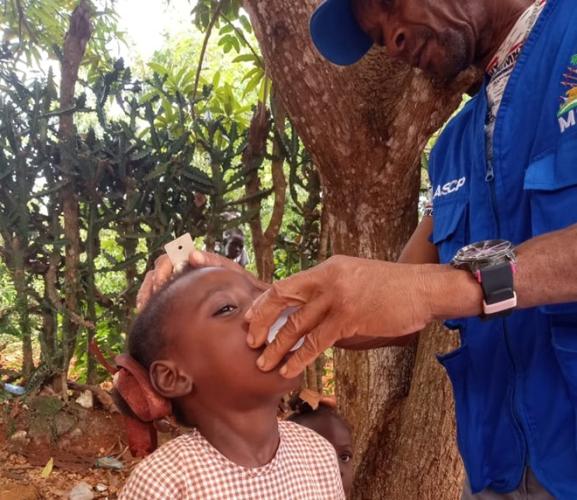 In the Haitian countryside, a man wearing a bright blue cap and a vest with official logos holds a small cup up to a young boy’s mouth.