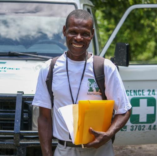 A Haitian man wearing a white T-shirt, backpack, and lanyard stands in front of a hospital vehicle, grinning. He holds papers and manila envelopes in his left hand.