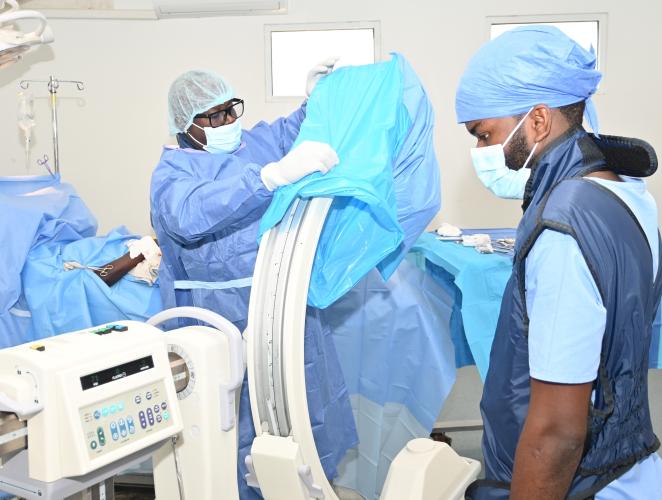 In a white operating room, two Haitian men in full PPE prepare for surgery. One arranges blue drapes over a C-arm device and the other checks a monitor.