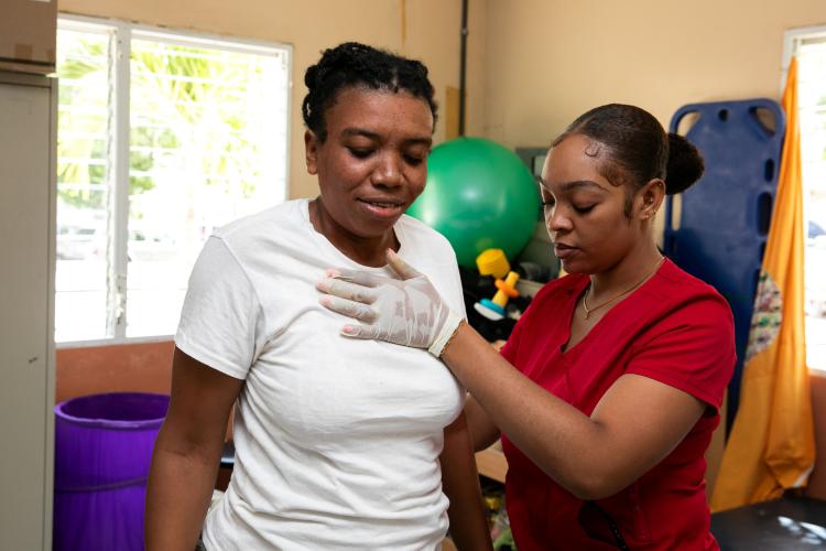 Two Haitian women, seen from the waist up, stand in a brightly-lit rehab center. One, in a red scrub top, presses a hand to the other woman’s upper chest, supporting her.