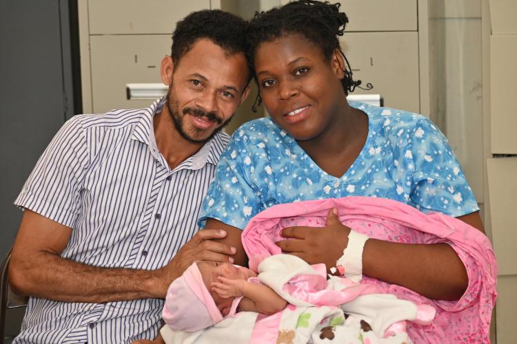 A light-skinned man with neatly-trimmed facial hair and a darker-skinned woman holding an infant in a pink blanket lean into each other, smiling happily.