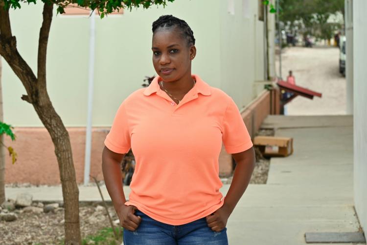 A Haitian woman community health worker stands in front of a building and gives a small smile at the camera. She wears a coral-colored polo shirt. Her hands are on her hips, and her hair is intricately braided.