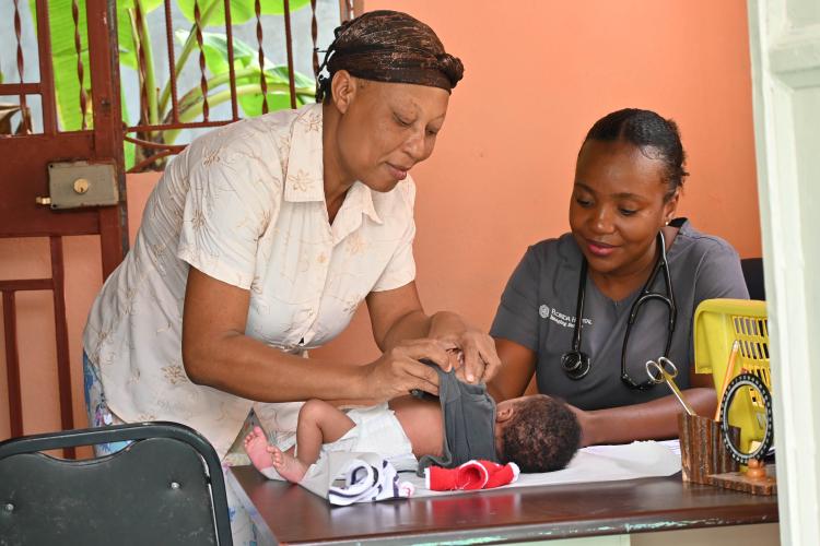 A Haitian woman leans over an examination table as she changes her baby. A Haitian woman doctor is sitting behind the table and is smiling at the baby.