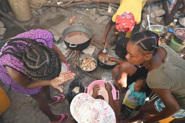 A group of Haitian women crowd around pots and pans to cook a mid-day meal for their children.