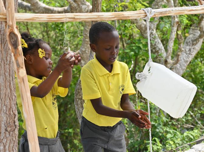 A young boy and girl in yellow school polo shirts wash their hands using a Tippy Tap device.