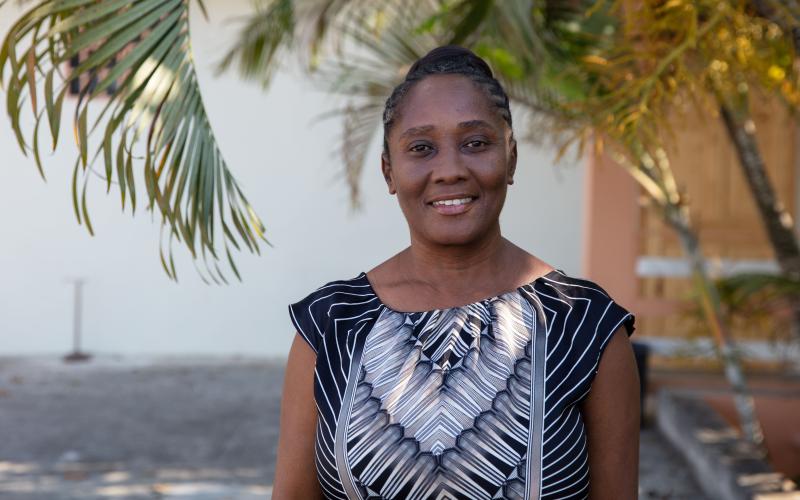 Headshot of a Haitian woman smiling, standing tall under palm fronds. Her hair is tightly braided and pulled back and she wears a black and white sleeveless dress.