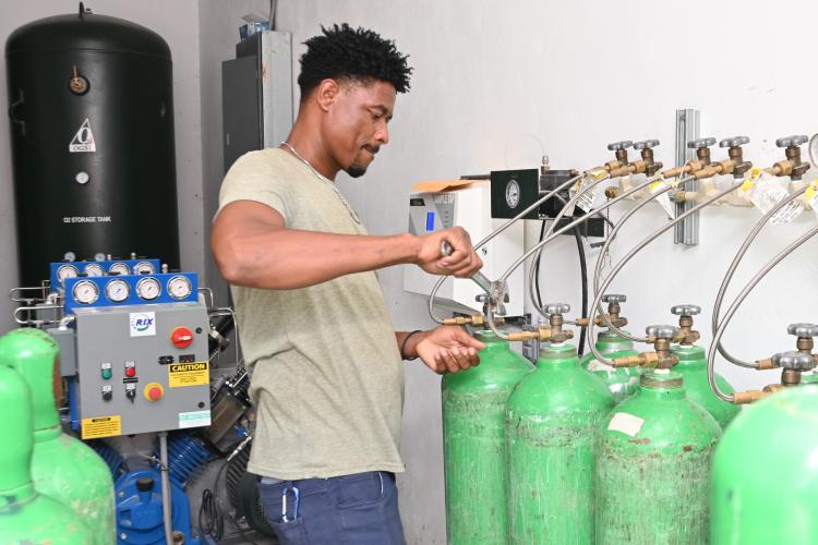 A Haitian man uses a large wrench to remove a green oxygen cylinder from an array of cylinders and hoses. On his left are blue compressors and a large black tank.