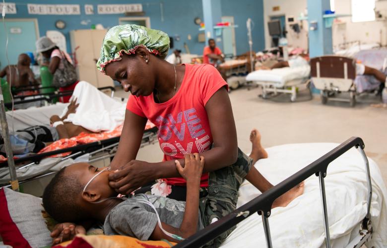 A woman in a red T-shirt that says “Love” sits with a young boy on a hospital bed, gently supporting his head. He curls into her, holding her arm.