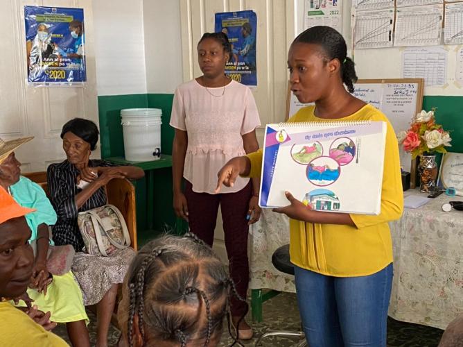 In a room with posters and charts on the walls, two Haitian women present information to a group of older Haitian women seated on chairs.