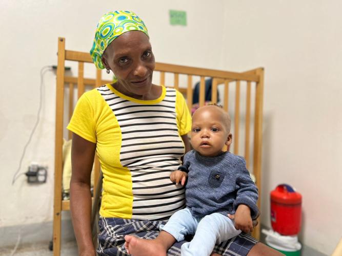 An elderly Haitian woman sits with a baby in her lap. The woman and baby sit in a chair in front of a wooden crib. The woman wears a green printed bandana over her head and a yellow short-sleeved top that has black and white stripes down the front. The baby wears a dark blue sweater and light blue pants.