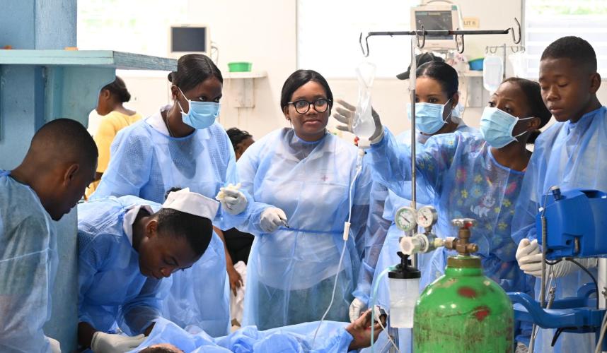 A half-dozen Haitian clinicians in blue medical gowns surround the bed of an ER patient who is receiving IV fluids.