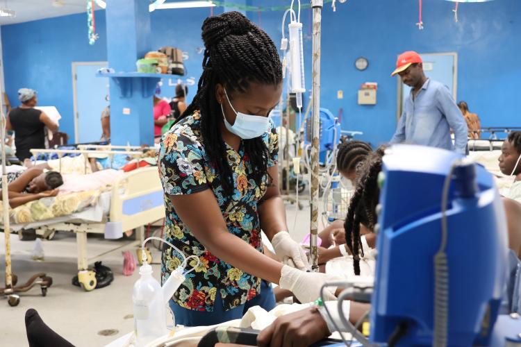 An woman Haitian emergency room clinician tends to the patient. The patient is mostly hidden behind a blue patient monitor. The clinician has long braids and wears a patterend scrub top, a blue mask and white exam gloves.