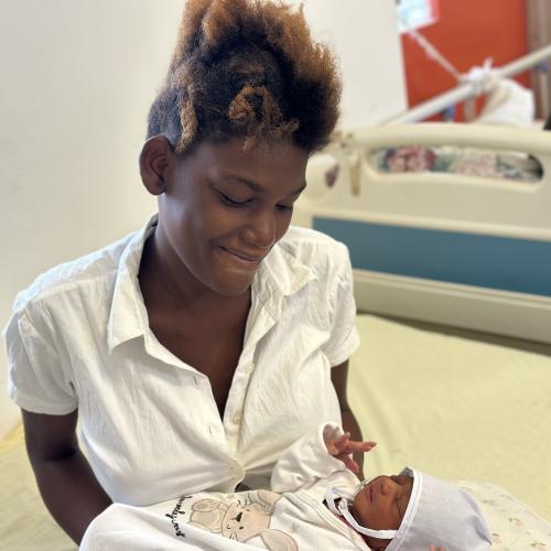 A young Haitian woman in a white blouse smiles down at the infant she holds in her arms.