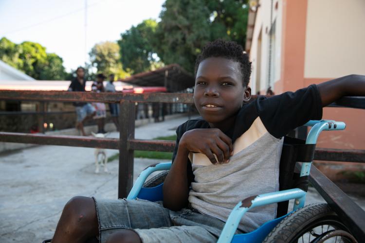 A Haitian teen sits in a bright blue wheelchair. His upper body is turned to face the camera. He is smiling.