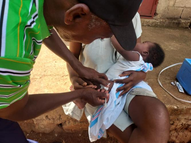 A male community health worker vaccinates a baby, who sits in his mother's lap.