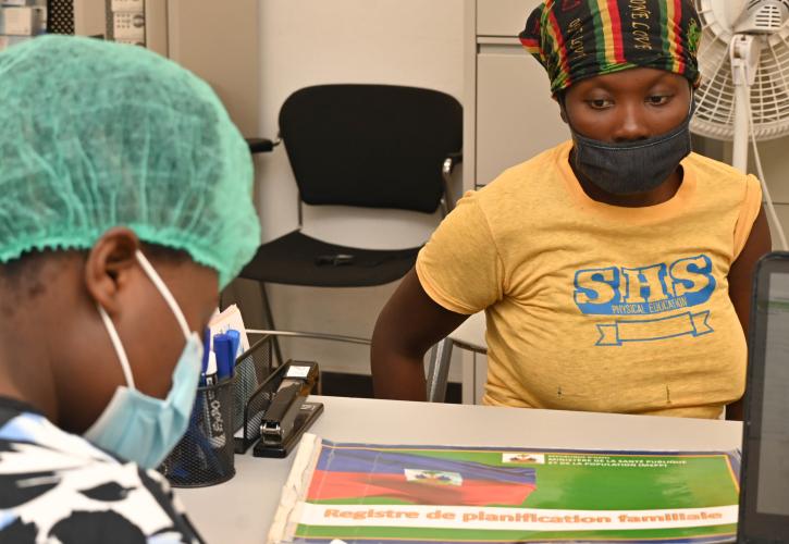 A woman in gold T-shirt, black/green/gold/red head scarf, and face mask sits facing a desk with office supplies and an oversize book with an image of the Haitian flag on its cover.