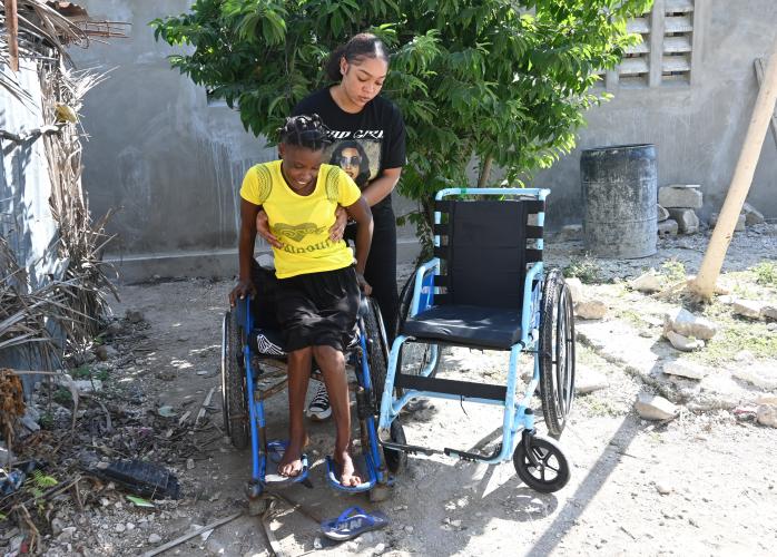 In a courtyard, one Haitian woman supports another as she lifts herself up from an old wheelchair, smiling. 