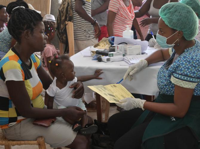 A Haitian woman holding a baby on her lap speaks with a nurse in face mask and gloves. They sit at a table, apart from a crowd.