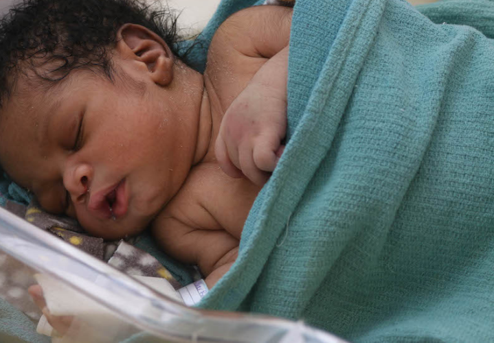 A Haitian baby rests in a bassinet in HEI/SBH's NICU. The baby has dark curly hair and is cuddled into a light blue blanket.