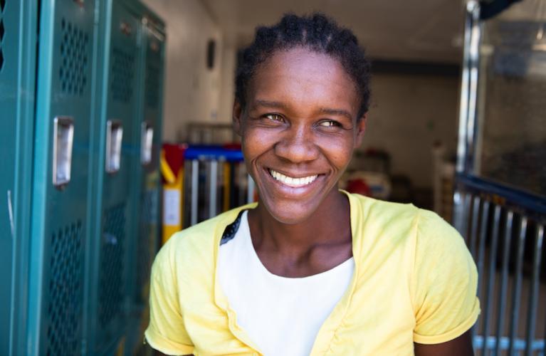 A Haitian woman shirt smiles in a doorway. She is shown from the shoulders up and wears a white t-shirt and a yellow cardigan. Her hair is braided.