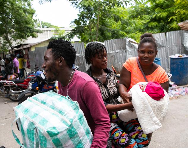 On a street corner in Haiti, two women hold an infant wrapped in a blanket. A man carrying a large tote bag passes next to them.