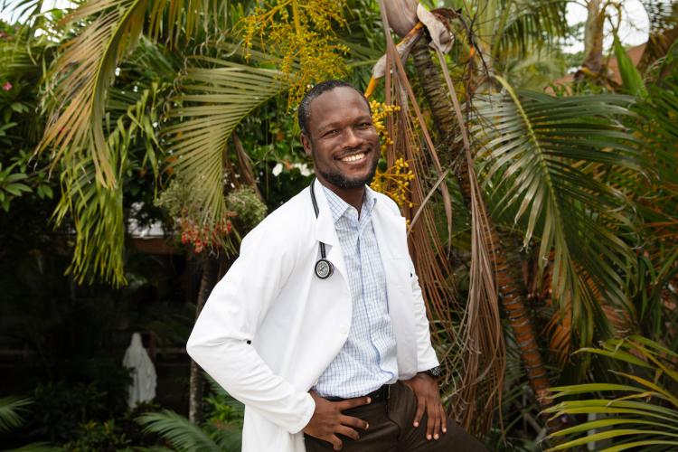 A Haitian male doctor stands in front of palm trees, smiling brightly at the camera. He wears a white doctor's coat and has a stethoscope around his neck.