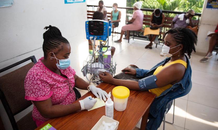 A nurse in pink scrubs and a blue medical mask checks the blood pressure of a woman patient during triage.