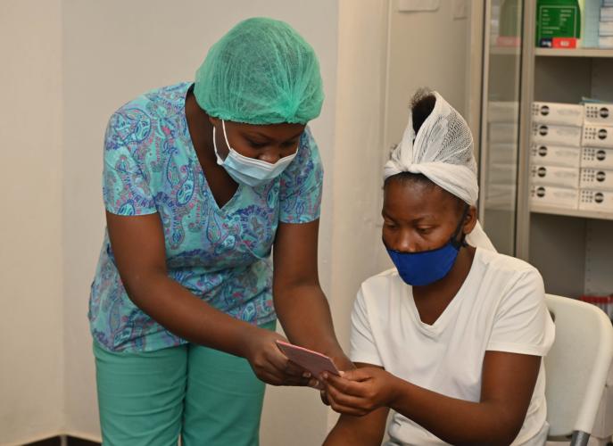 A Haitian woman in bright aqua scrubs and face mask smiles as she hands a pink card to another Haitian woman wearing a white shirt and head wrap and blue face mask.