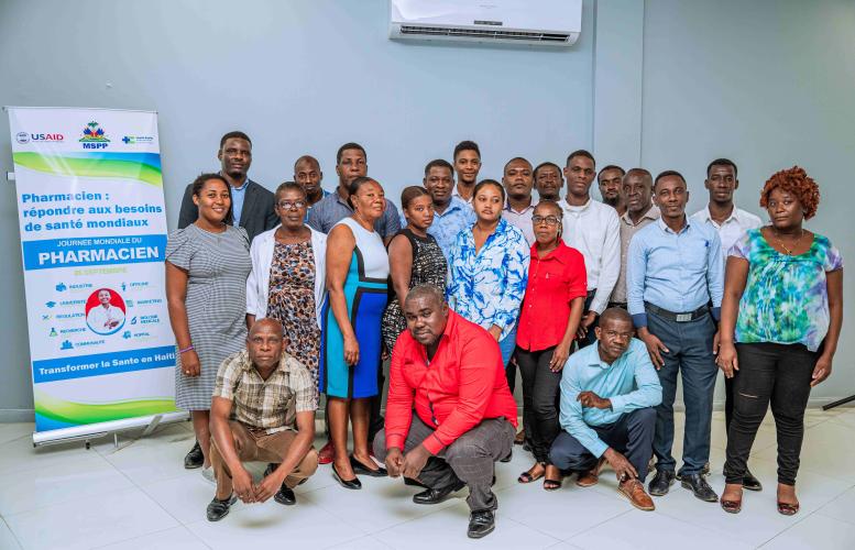 22 Haitian men and women, in business casual attire, pose for a group photo next to a World Pharmacists Day banner, in French, with USAID, MSPP, and HEI logos.