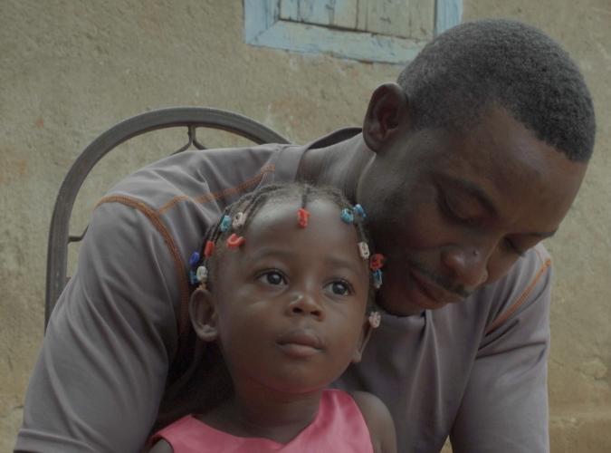 Close-up of a toddler in shiny pink dress and barrettes, gazing into the distance while sitting on the lap of her father who bends over her tenderly.