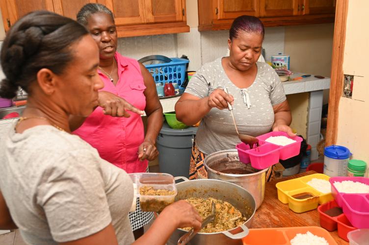 Three Haitian women stand around a kitchen table, spooning white rice, black bean sauce, and mixed vegetables into colorful single-serving containers.