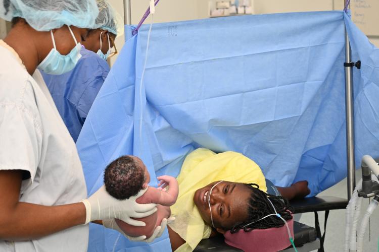 In an operating room, a nurse in full PPE holds out an infant to her joyful mother who is still on the operating table, under drapes.