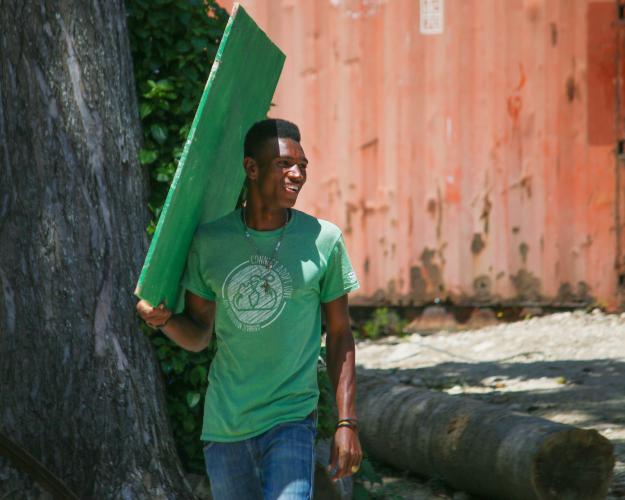 A Haitian man wearing a green T-shirt and blue jeans looks over his shoulder and smiles. He is carrying a piece of green-painted plywood.
