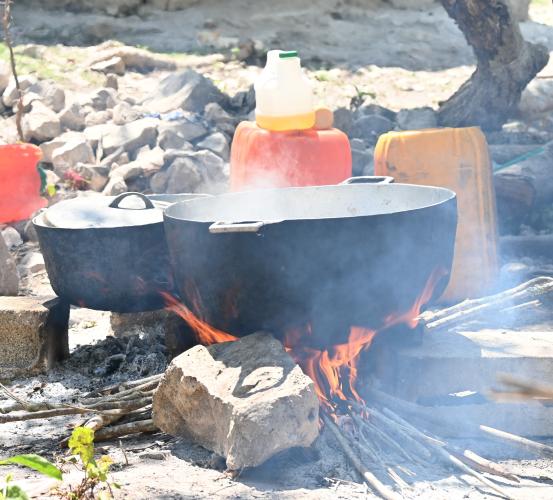 Two large pots sit on cinderblocks and rocks above an open fire in a sunny yard. One pot is covered, and the other has steam rising from it.